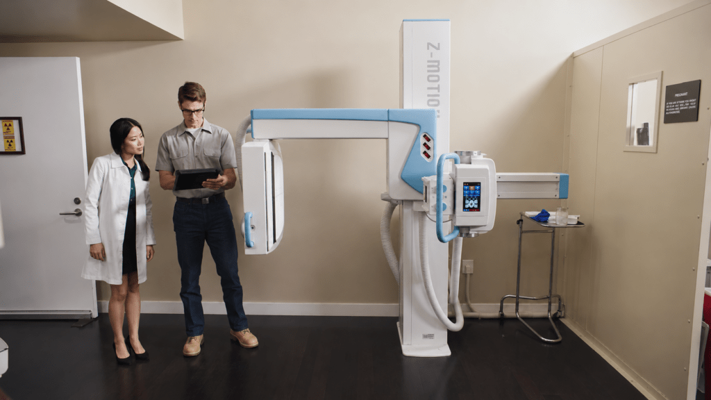 Two people standing beside a wall-mounted X-ray system in an imaging room